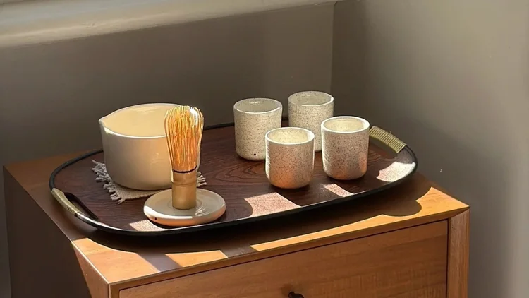 Ceramic cups and a matcha whisk on a tray atop a timber side table